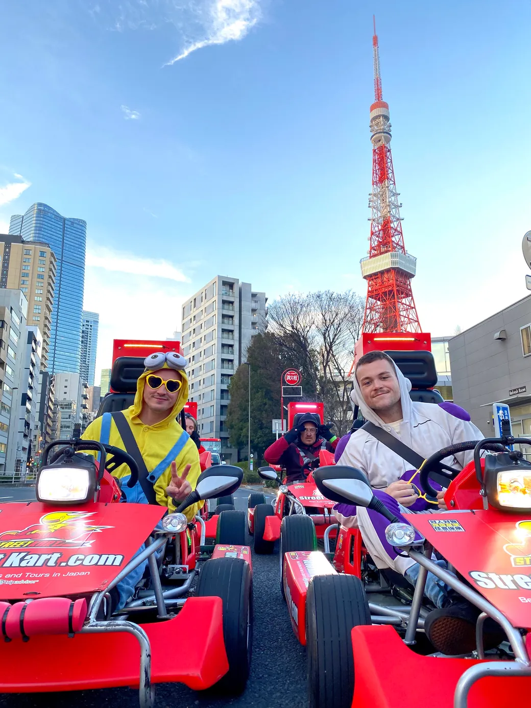 Group of people in red go-karts wearing colorful costumes on a city street with Tokyo Tower in the background, sunny sky above.