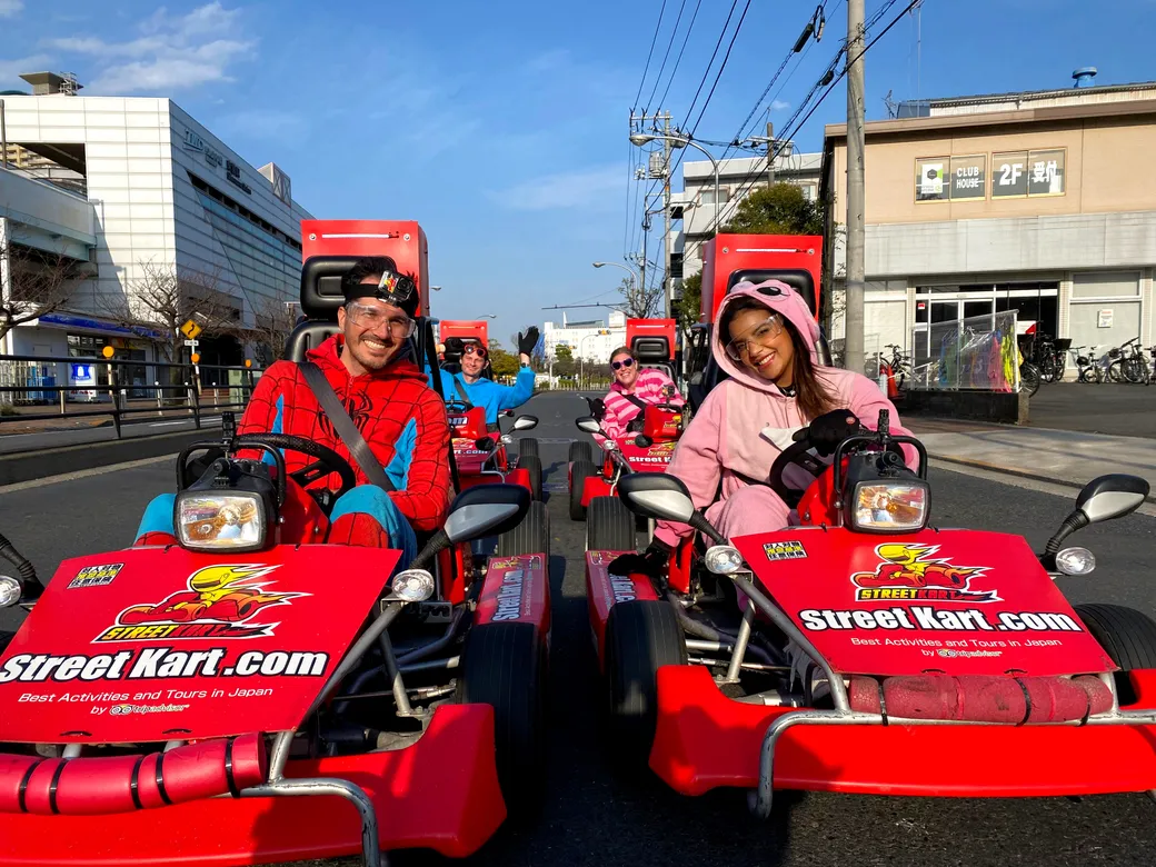 People in red Street Kart go-karts racing down a city street; two drivers in costume (Spider-Man and pink hoodie) smile at camera with others behind them.