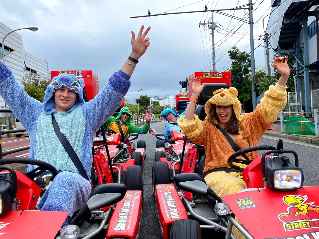 A group of people in animal onesies cheer and wave from red go-karts on a city street during a fun event.
